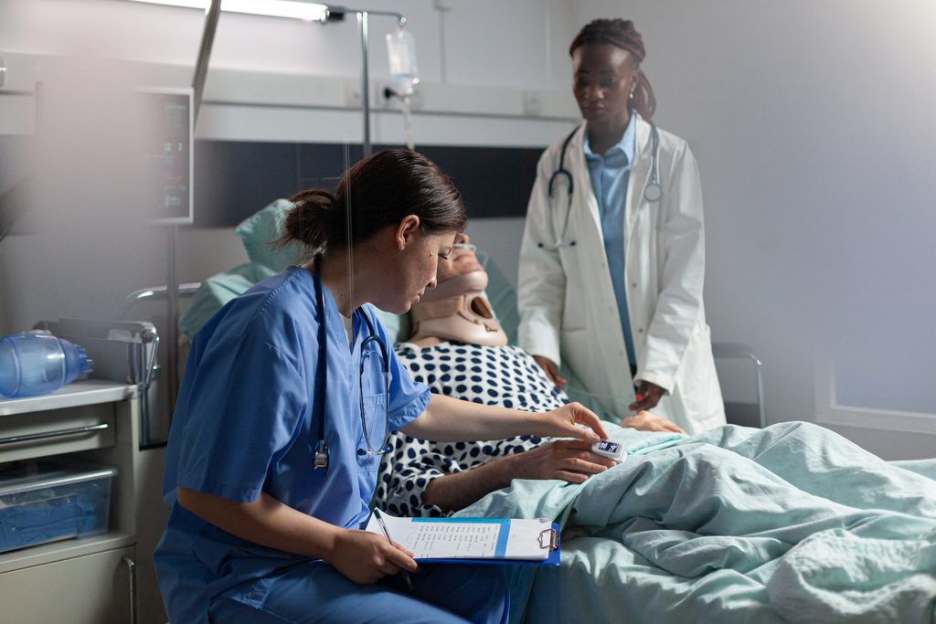 ICU nurse in blue scrubs checks a pulse oximeter reading on a bedbound patient wearing a cervical collar while a doctor in a white coat observes at the bedside.